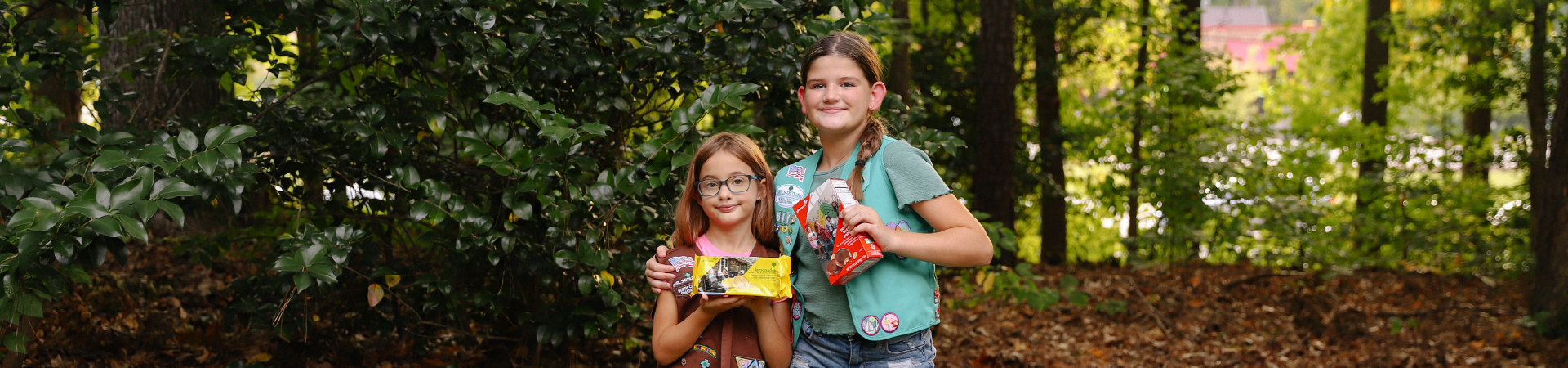  Two Girl Scouts holding boxes of cookies near a forest 