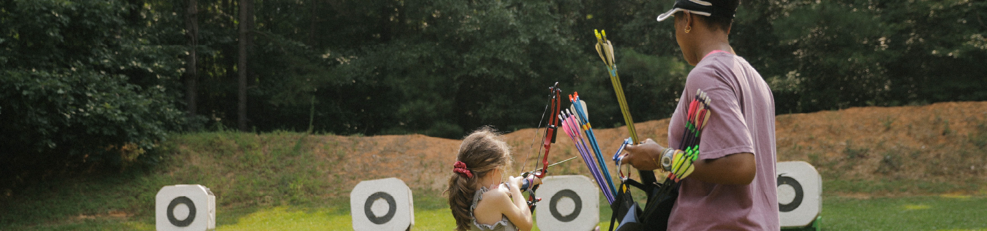  women in mauve shirt holding archery arrows for young girl shooting archery 